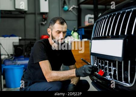 Close up photo of young male worker in black protective gloves cleaning radiator grille in foam with special brush. Carwash and detailing. Washing Stock Photo