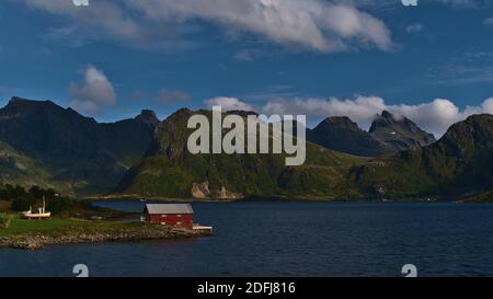 Red painted small shed on the shore of fjord Selfjorden on Moskenesøya island, Lofoten, Norway with majestic mountains of Flakstadøya in background. Stock Photo