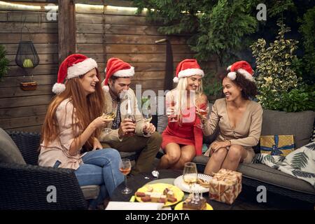 small group of friends having a party for christmas, drinking alcohol, talking, laughing, sitting outdoor in the backyard on the sofa Stock Photo
