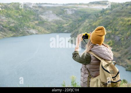 Rear view of young female backpacker in warm casualwear taking photo of riverside or lake surrounded by mountains during weekend trip Stock Photo