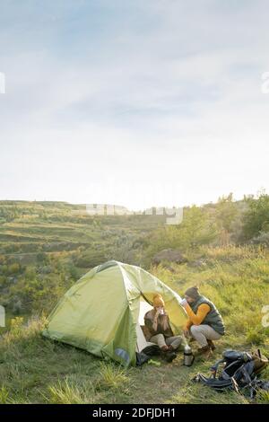 Young man squatting on green grass by tent where his wife relaxing and both having hot tea after long way during trip on autumn weekend Stock Photo