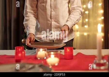 Hands of young man in white shirt going to put plates on festive table decorated with burning candles for romantic dinner with his wife Stock Photo