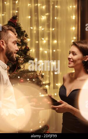 Happy elegant young woman giving red toy balls to her husband while looking at him with smile against Christmas tree in home environemnt Stock Photo
