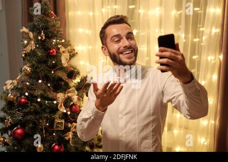 Young cheerful bearded man in white shirt looking at smartphone screen while making selfie or communicating to someone against Christmas tree Stock Photo