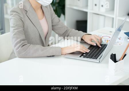 Hands of young contemporary businesswoman in formalwear and protective mask pressing keys of laptop keypad during network Stock Photo