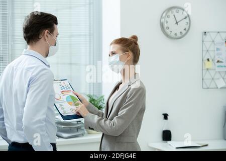 Two young elegant accountants in protective masks discussing financial data in document with diagrams and charts at meeting in office Stock Photo