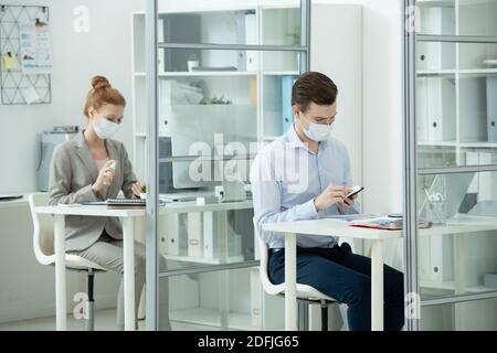 Young businessman in protective mask and formalwear using smartphone while his female colleague spraying sanitizer on hands behind him Stock Photo