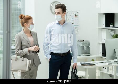 Two young managers in protective masks and formalwear discussing after work plans while leaving office and going to spend time together Stock Photo