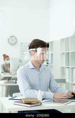 Young businessman in protective screen on head and formalwear looking at laptop display while working with data against female colleague Stock Photo