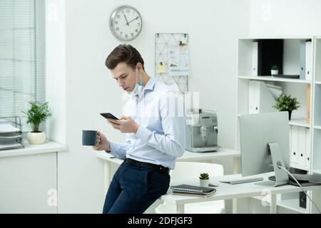 Young businessman in protective mask and formalwear using smartphone while sitting on desk with office supplies and having tea or coffee Stock Photo