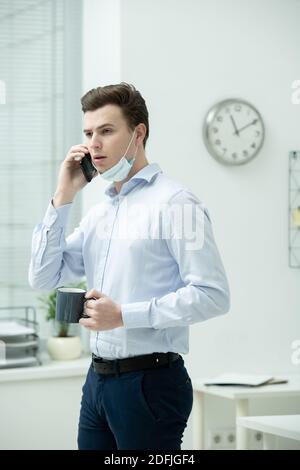 Young businessman in protective mask and formalwear consulting client on smartphone while standing by desk and having tea or coffee Stock Photo