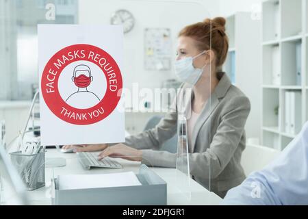Young contemporary businesswoman using laptop by desk in front of paper with recommendations about wearing protective masks Stock Photo