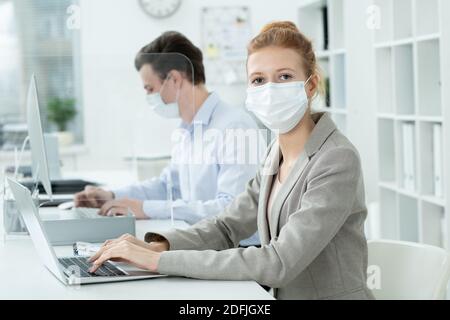 Young businesswoman in protective mask and elegant grey suit looking at you on background of her male co-worker analyzing online data Stock Photo