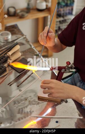 Hands of contemporary female artisan sitting by workplace and burning stick shaped workpiece with fire while holding it over burner Stock Photo