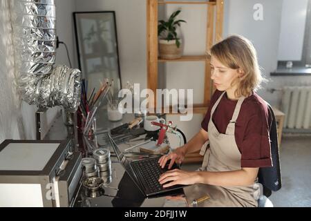 Serious young blond craftswoman in apron touching buttons of laptop keypad while sitting by table in workshop or studio and entering data Stock Photo