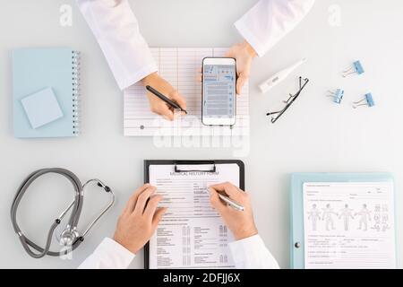 Overview of hands of male clinician filling in medical history form and his assistant making notes on blank card and scrolling in smartphone Stock Photo
