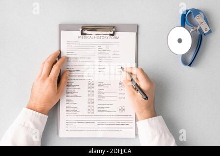 Overview of doctor hands with pen over medical history form in clipboard filling in personal information of patient with clinic tool near by Stock Photo