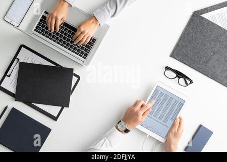 Flatlay of hands of contemporary economist using tablet while analyzing financial data while his colleague pressing buttons of laptop keypad Stock Photo