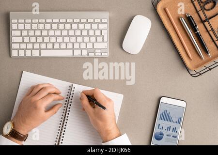 Flatlay of hands of young male broker with pen over blank page of open notebook making notes among office supplies, mouse, gadget and keypad Stock Photo