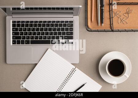 Top view of cup of coffee, open notebook with pen and blank pages, laptop and basket with clips and other office supplies on workplace Stock Photo