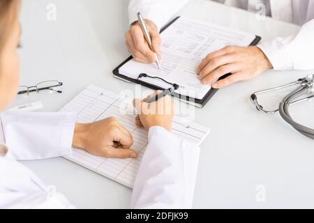 Hands of young female clinician with pen filling in blank cards while sitting in front of her male colleague putting information in medical document Stock Photo