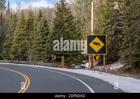 A large moose crossing sign with a moose icon inside a yellow diamond shape warns motorists of the hazard of crossing moose. The warning stands on the Stock Photo