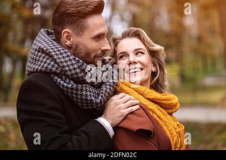 Close up. Happy in love young people, man hugging woman from behind when she looks at him, happy couple walking in a autumn park wearing stylish coats Stock Photo