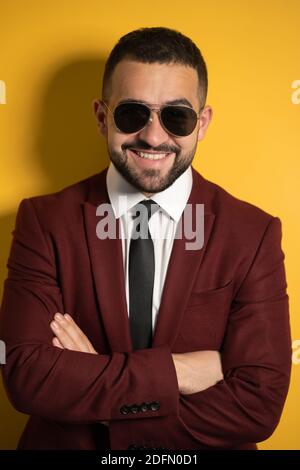 Close up portrait of handsome business man in sunglasses and burgundy suit looking at camera with hands folded isolated on yellow background Stock Photo