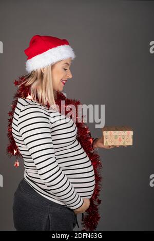 Beautiful pregnant woman in third trimester is wearing Santa hat and red christmas garland on grey background, holding a gift box Stock Photo