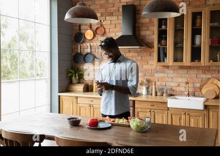 Young happy african american man using smartphone, distracted from cooking. Stock Photo