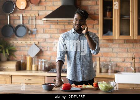 Smiling young mixed race man answering phone call, preparing food. Stock Photo