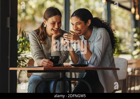 Two beautiful women sitting in a coffee shop talking to each other and smiling. Women in conversation while meeting up for coffee. Stock Photo