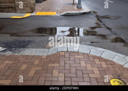 Round cornered brick and stone sidewalk alongside asphalt street with puddles and wet with rain, horizontal aspect Stock Photo