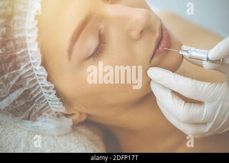 Beautician doing beauty procedure with syringe to face of young brunette woman in sunny clinic. Cosmetic medicine and surgery, beauty injections Stock Photo