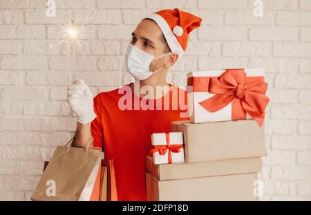Young man courier in medical face mask, protective gloves, santa hat deliver Christmas, New Year presents. Happy delivery man hold sparkler light, sho Stock Photo