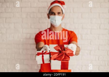 Happy man courier in santa hat, medical face mask, protective gloves give Christmas New Year presents and smile. Young delivery man in face mask carry Stock Photo