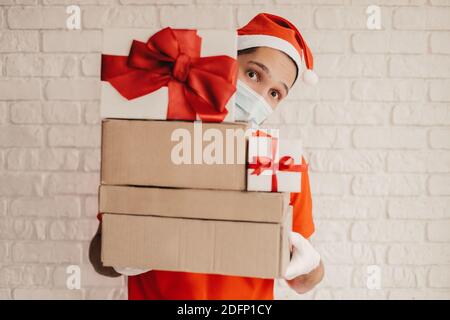 Happy funny festive delivery man in santa hat, medical face mask, protective mask carry Christmas New Year presents. Young man courier in face mask lo Stock Photo
