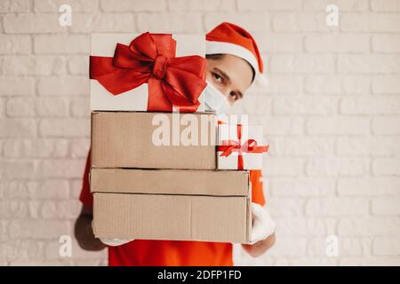 Young happy man courier in protective gloves, Santa hat, medical face mask carry New Year Christmas presents, smile. Funny delivery man in face mask l Stock Photo