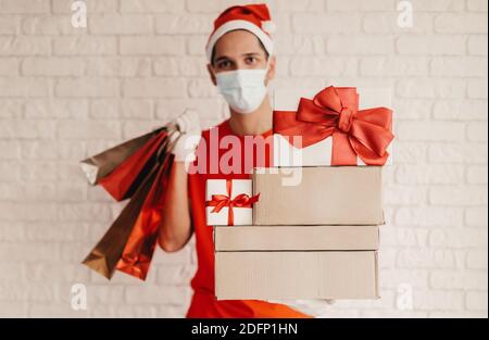 Young man courier in protective face mask, Santa hat, medical glove deliver Christmas presents. Happy delivery man in face mask carry shopping bags, c Stock Photo