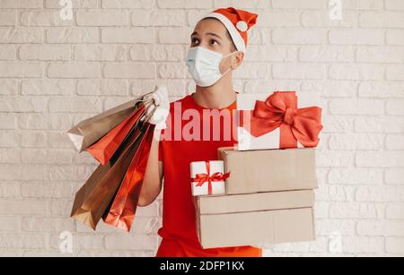 Happy man courier in Santa hat, medical face mask, protective gloves deliver Christmas presents. Surprised delivery man in face mask carry shopping ba Stock Photo