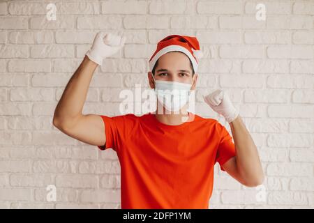 Young positive delivery man in santa hat, medical face mask, protective gloves celebrate success, win. Happy man courier in protective mask cheer with Stock Photo
