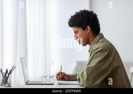 Side view of millennial black student sitting at desk with laptop, making notes in copybook, learning remotely from home Stock Photo