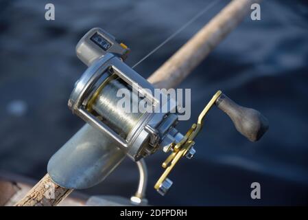 Round silver fishing reel with a line counter indicating almost 10 meters fishing line out place in a rod holder with blurred river water background. Stock Photo