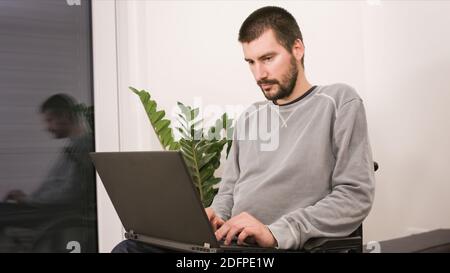 Young man in wheelchair with laptop working remotely from home, using internet and looking at screen. Stock Photo