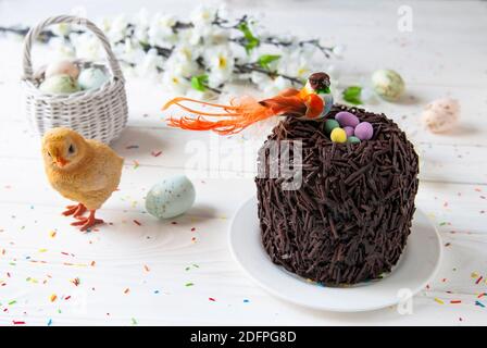 Easter chocolate cake and bird sitting in the nest. Little orange bird, colored eggs, chick and wicker basket on a background of Easter’s décor Stock Photo