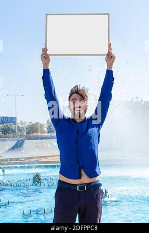 Young bearded handsome smiling man dressed in blue casual wear with an inviting gesture, holding wood framed blank whiteboard in blue sky background. Stock Photo
