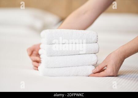 Close up of female hands putting stack of fresh white bath towels on the bed sheet in room. Hotel service concept Stock Photo