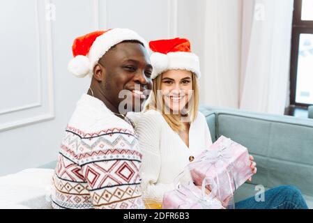 A young couple in love is sitting on the sofa and exchanging gifts. Boyfriend and girlfriend. Beautiful young couple is celebrating at home. Stock Photo