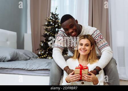 Beautiful young couple is celebrating Christmas and New year at home. Romantic multiethnic couple. Beautiful woman is happy with the gift, smiles and Stock Photo