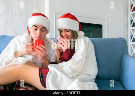 Young Caucasian couple drinking hot tea or cocoa while sitting at home on the sofa wearing santa hats and pajamas wrapped in a white blanket Stock Photo